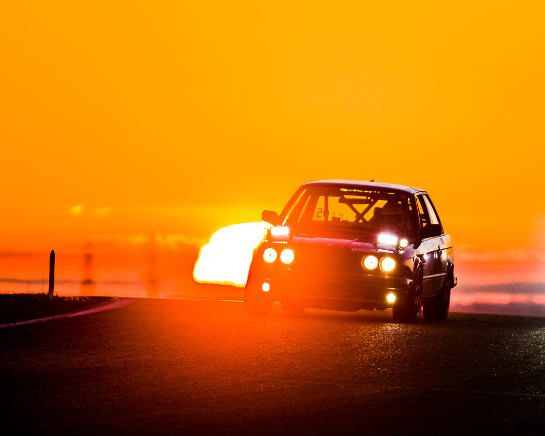 Photo of the Shift-Up Now BMW at Sunrise during the NASA 25 Hours of Thunderhill, headlights shining and a brilliant sun rising behind the car.